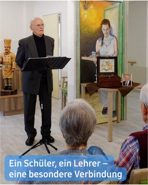 Ein älterer Mann in einem dunklen Anzug steht an einem Rednerpult und spricht oder singt. Er befindet sich im Vordergrund, leicht versetzt zur Mitte. Das Rednerpult ist schwarz und mit einer musikständerähnlichen Oberseite ausgestattet.  Hinter ihm ist eine gemalte Tür dargestellt, die eine jüngere Frau in einem hellen Kleid zeigt, die an einem Tisch mit einem Musikinstrument oder einer Spieluhr sitzt. Das Gemälde ist im Rahmen der Tür eingefasst und vermittelt den Eindruck einer Szene aus einer Erinnerung oder einer vergangenen Zeit. Ein gerahmtes Foto eines Mannes im Anzug ist auf einem Beistelltisch in der dargestellten Szene sichtbar.  Im Vordergrund, unten rechts und teilweise vom unteren Bildrand verborgen, sind die Rücken von zwei Personen zu sehen. Die Umgebung scheint ein Pflegeheim zu sein.  Eine Beschriftung am unteren Rand lautet: "Ein Schüler, ein Lehrer – eine besondere Verbindung."
